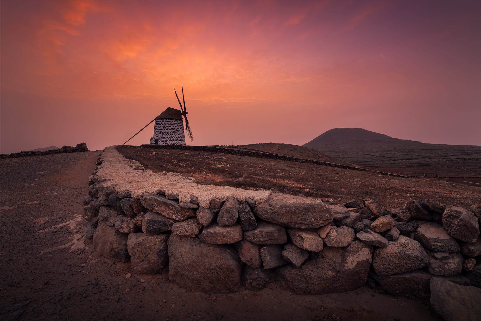 Traditional windmill in Villaverde at sunset, surrounded by the volcanic landscape of Fuerteventura beneath a glowing orange sky, Canary Islands, Spain.