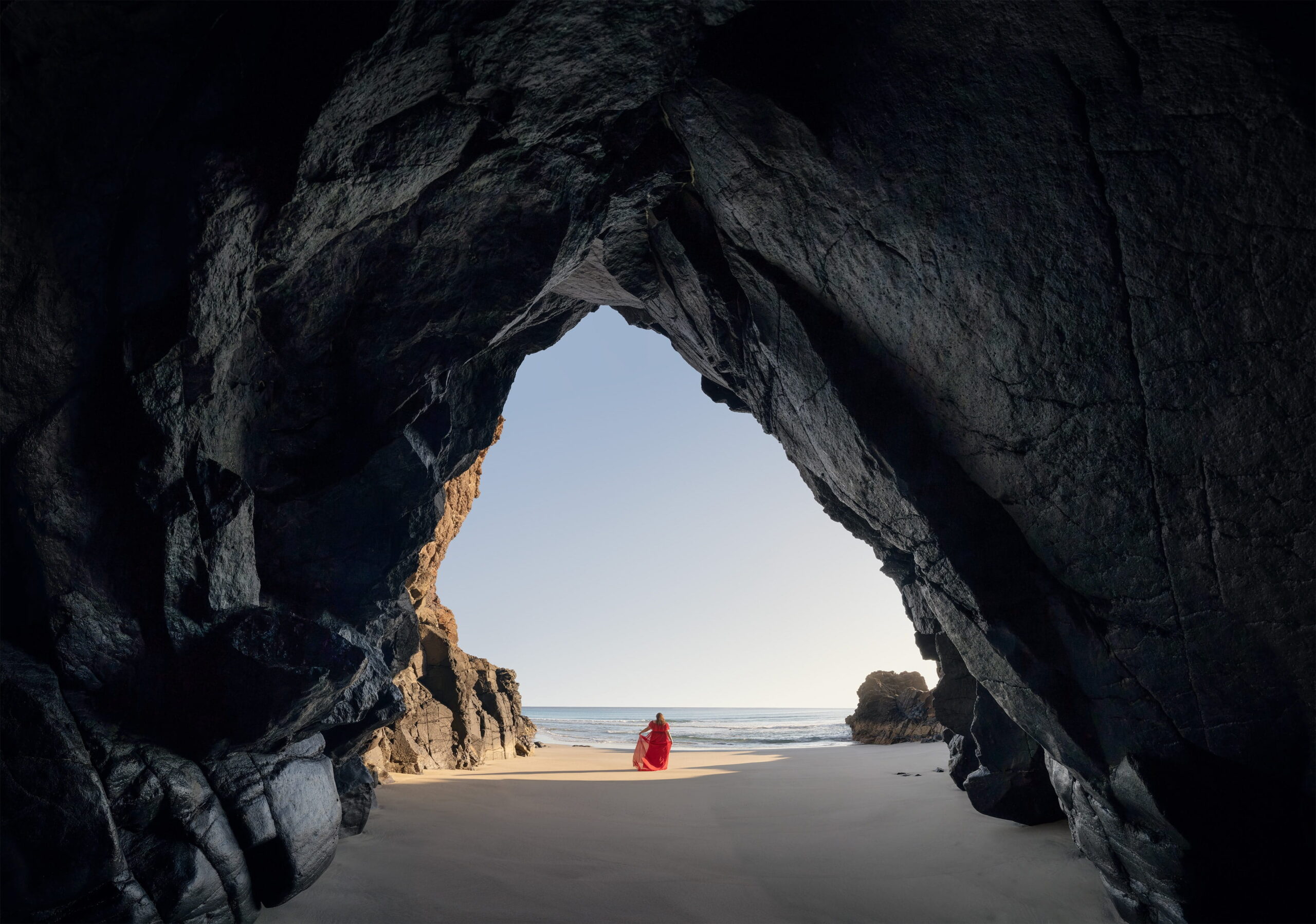 Panoramic landscape photograph inside the Cueva de Órzola in Lanzarote, Canary Islands, with a woman in a flowing red dress framed by the dark volcanic cave and the sunlit beach and Atlantic Ocean beyond.