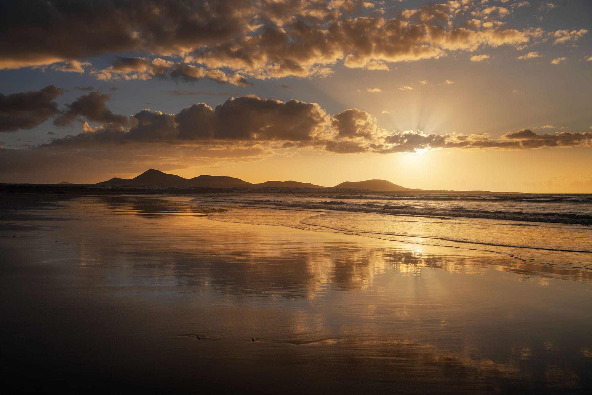 Golden sunset over Famara Beach in Lanzarote, Canary Islands, with dramatic clouds and sunlight reflecting across the wet sand and Atlantic Ocean.