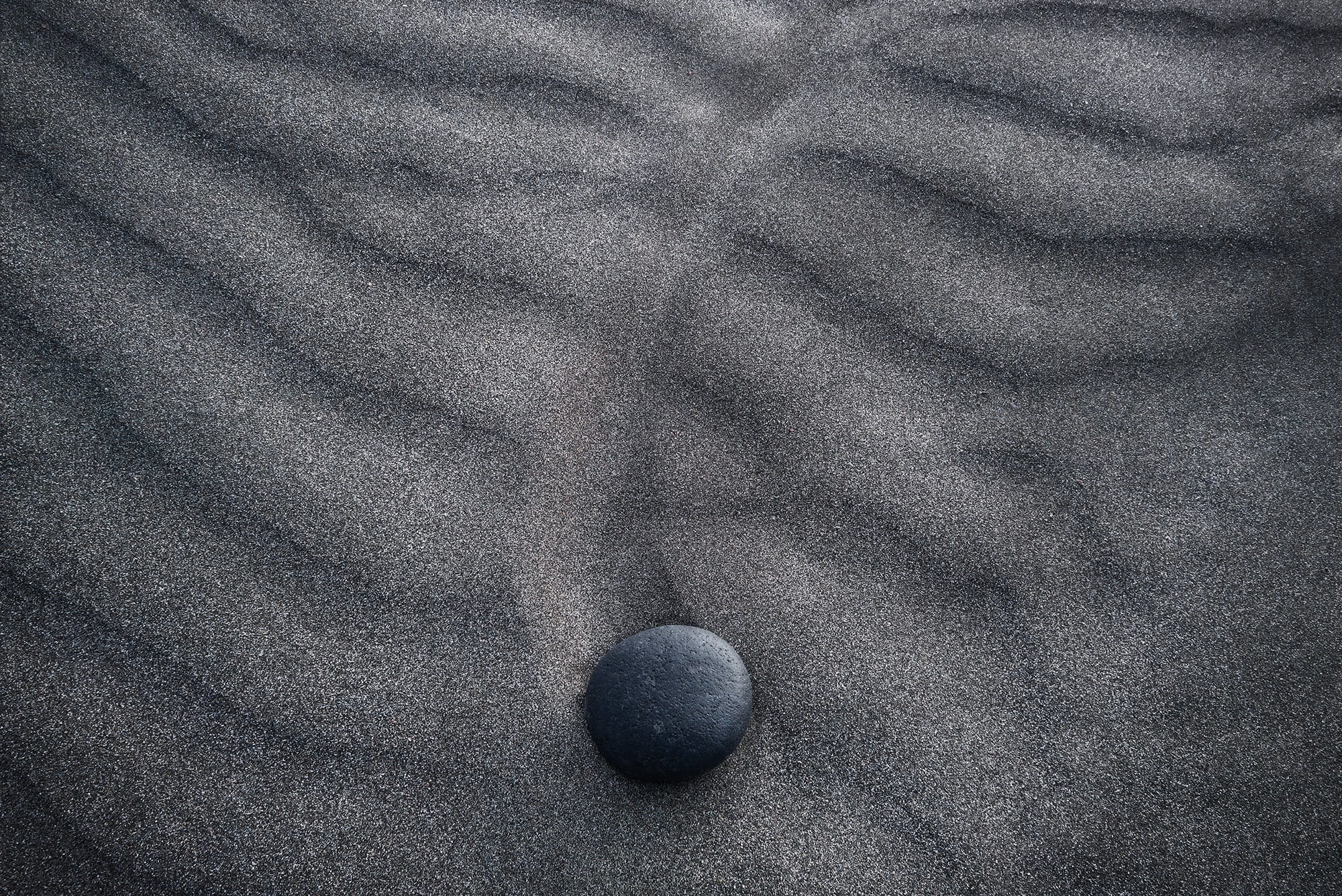 Minimalist landscape of a single volcanic stone resting on rippled black sand in Lanzarote, Canary Islands, Spain.