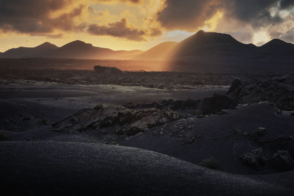 Golden sunset over the volcanic landscape of Timanfaya National Park in Lanzarote, Canary Islands, with sun rays illuminating black lava fields and silhouetted volcanic peaks.