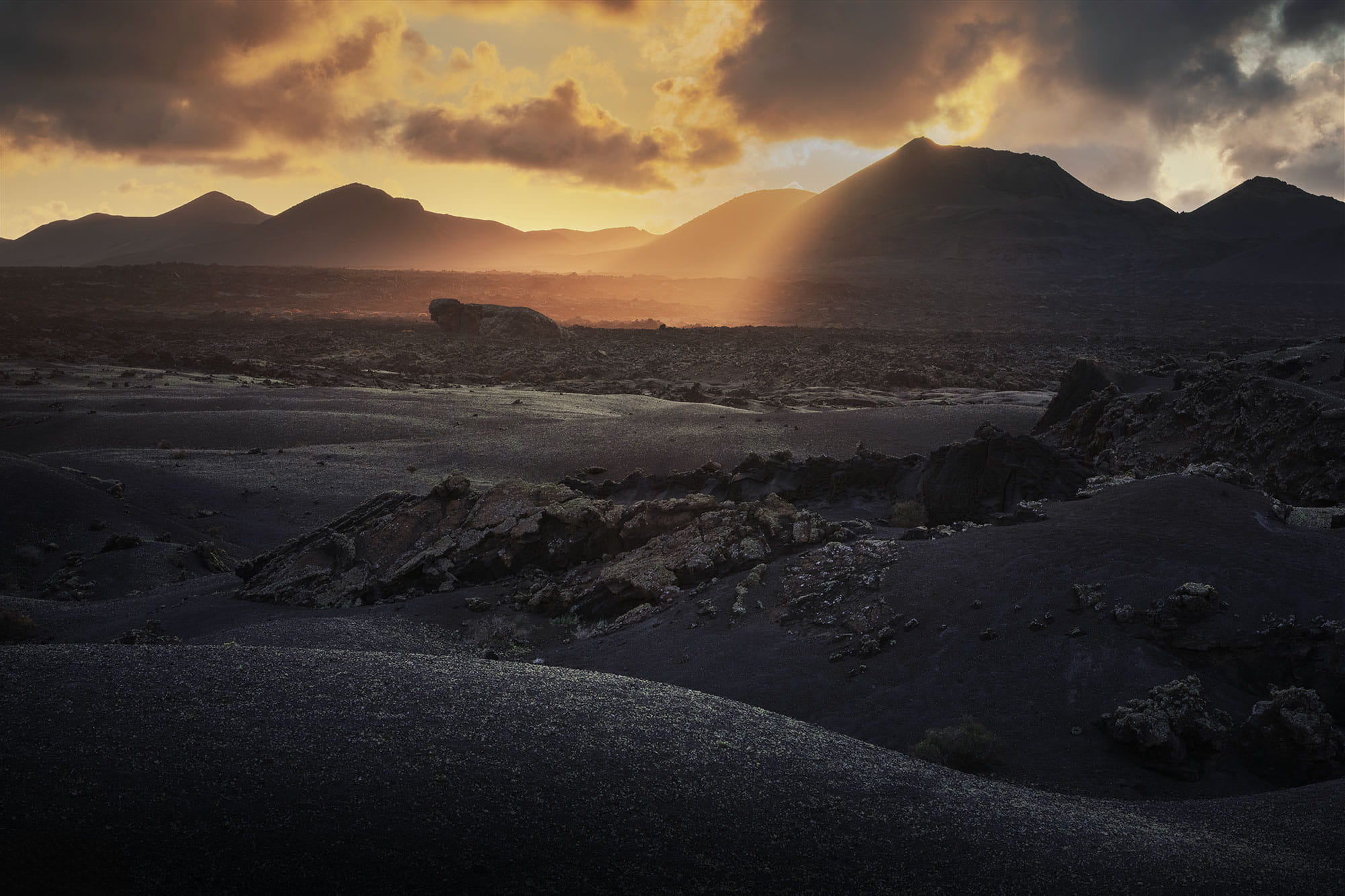 Golden sunset over the volcanic landscape of Timanfaya National Park in Lanzarote, Canary Islands, with sun rays illuminating black lava fields and silhouetted volcanic peaks.