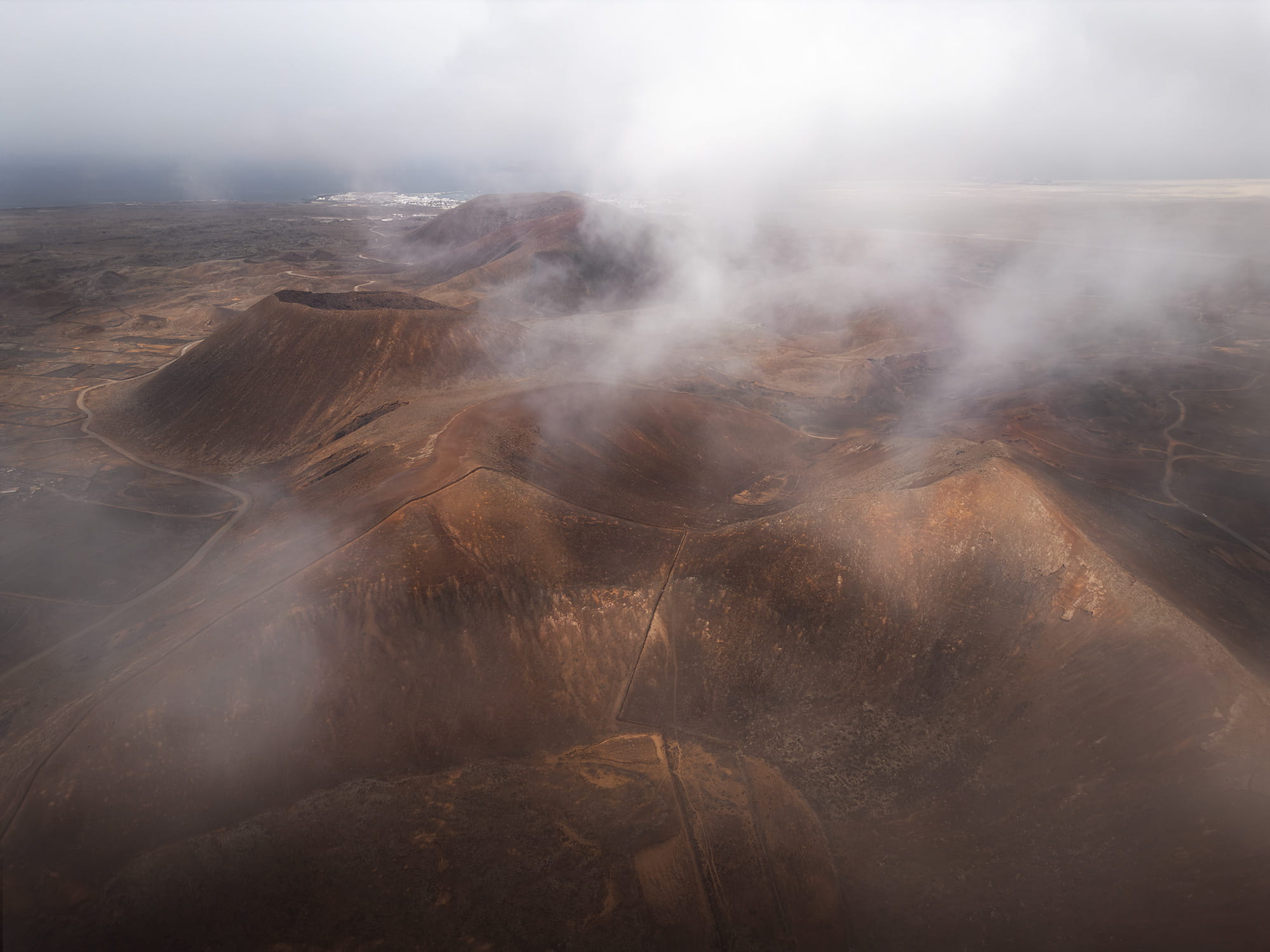 Aerial landscape photography of the Volcanes de Bayuyo in Fuerteventura, Canary Islands, featuring reddish-brown volcanic craters partially covered by drifting clouds, creating a mysterious atmosphere.