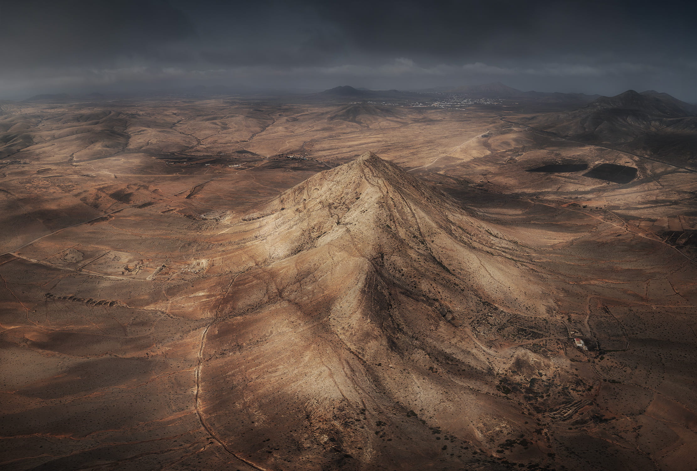 Aerial landscape photography of Tindaya Mountain rising from the arid volcanic landscape of Fuerteventura, Canary Islands, under dramatic cloudy skies.