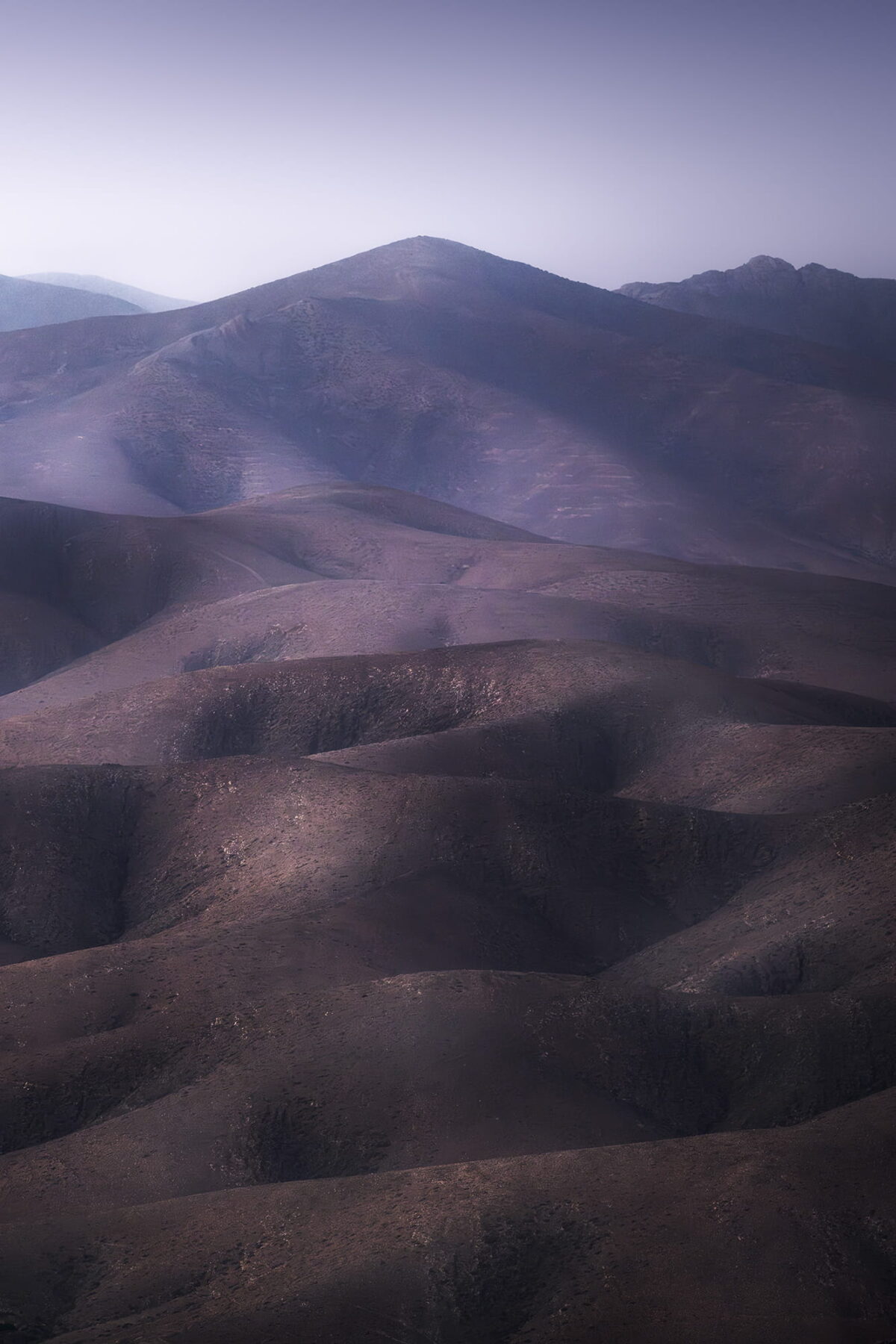 Landscape photography of layered volcanic hills in Fuerteventura, Canary Islands, featuring soft purple tones, gentle curves and distant mountain peaks fading into the hazy evening light.