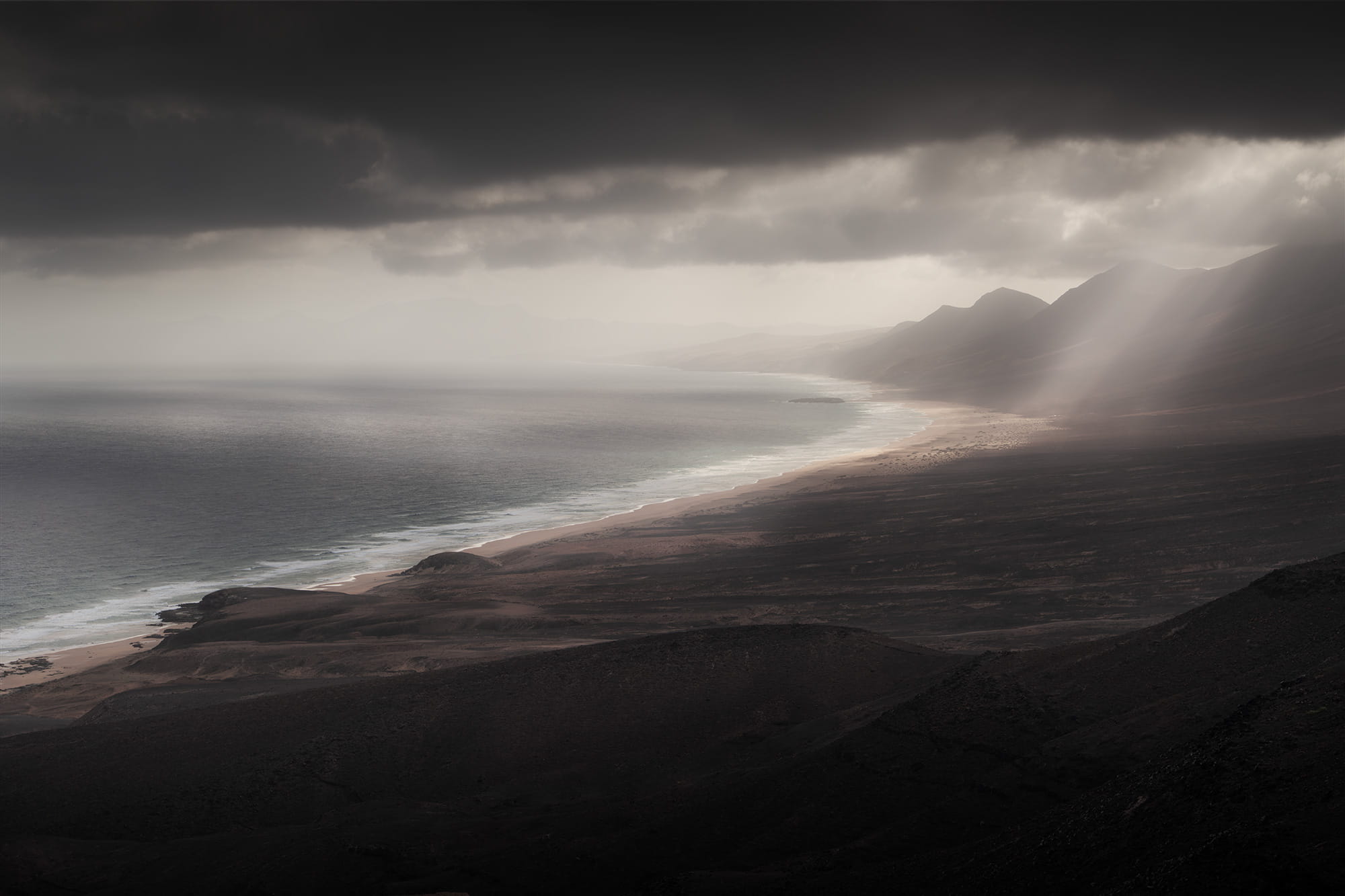 Panoramic landscape photography of Cofete Beach from the Cofete viewpoint in Fuerteventura, Canary Islands, featuring a wild coastline, rugged mountains and dramatic sun rays breaking through dark storm clouds.