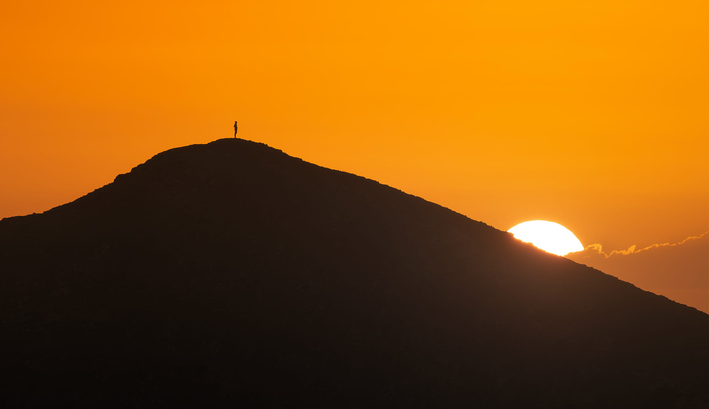 Minimalist landscape photography of a lone person standing on the silhouette of the Bayuyo volcanic ridge in Fuerteventura, Canary Islands, against a vibrant orange sky as the sun sets behind the mountain.