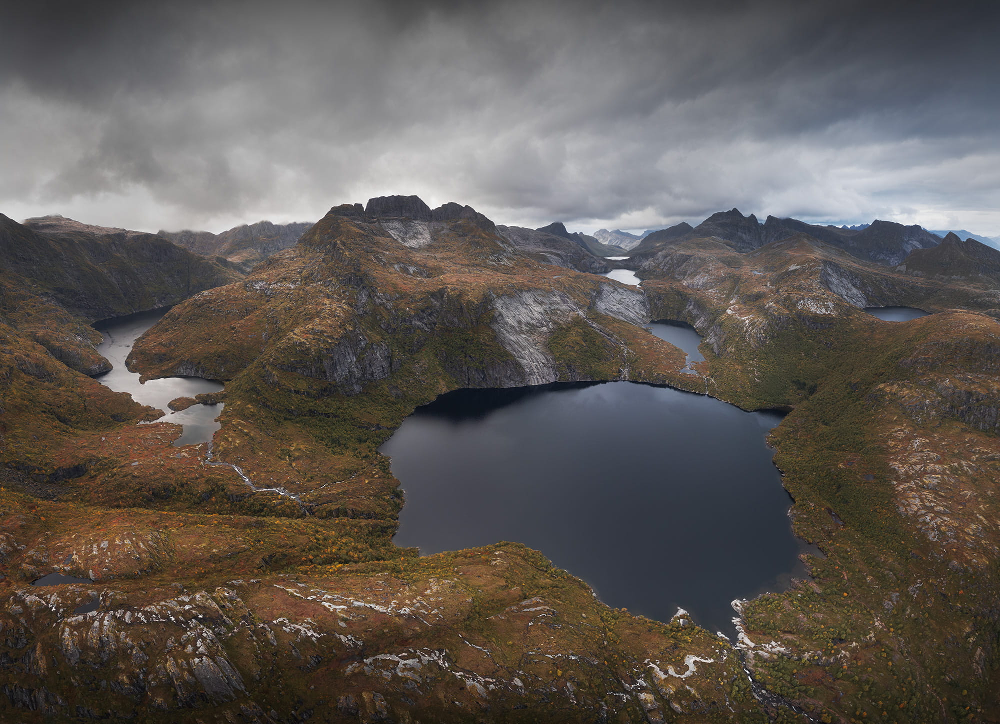 Aerial panoramic landscape photography of Sørvågen in the Lofoten Islands, Norway, featuring dramatic mountains, dark alpine lakes and colorful autumn vegetation beneath a moody, stormy sky.
